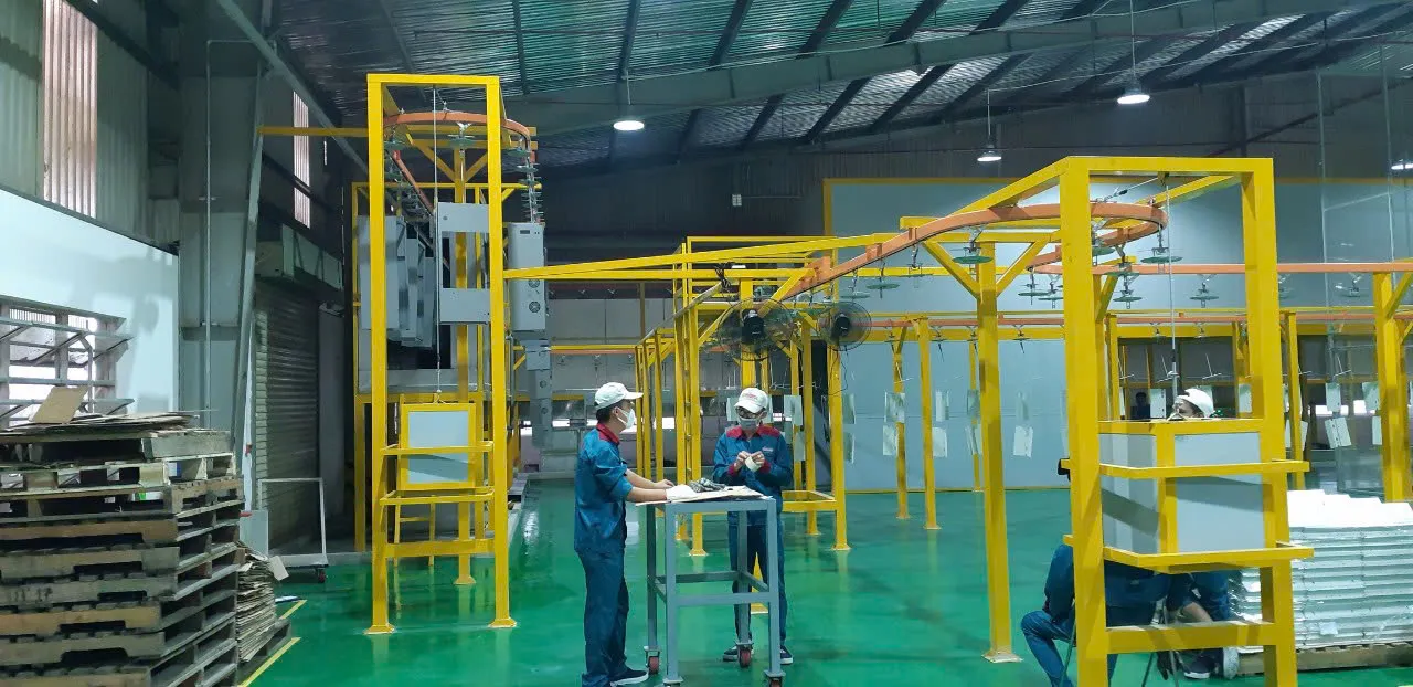 Dây chuyền sơn tĩnh điện tự động An Khanh lắp đặt với khung xích tải màu vàng, sản phẩm dạng tấm treo trên móc và nhân viên chuẩn bị hàng tại khu vực treo